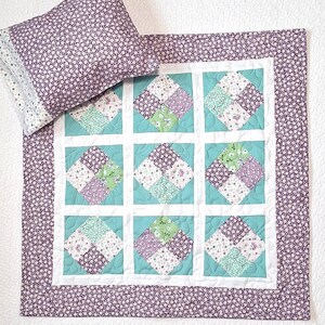 May include: A quilted baby blanket and pillow set. The blanket features a grid pattern with teal squares and floral patchwork. The border and pillow are covered in a small purple floral print. The set is designed for a nursery.