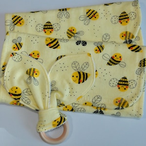 May include: A yellow baby blanket and teether set with a repeating pattern of smiling bees. The teether has a wooden ring and a soft, textured fabric with bee-shaped ears. The blanket is folded and features the same bee design.