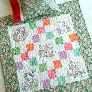 May include: A white baby quilt with a patchwork design featuring embroidered animals. The quilt has a green floral border and a matching pillow.