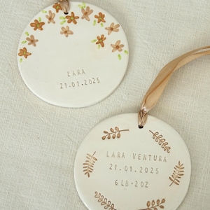 May include: Two white ceramic ornaments with tan ribbons. One has brown flowers and the name "LARA" and the date "21.01.2025". The other has the name "LARA VENTURA", the date "21.01.2025", and the weight "6 lb 2 oz".