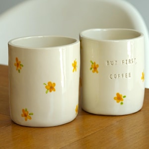 May include: Two cylindrical off-white ceramic mugs featuring a hand-painted pattern of small yellow flowers with green stems. One mug displays the embossed text "BUT FIRST, COFFEE" in capital letters on the front, while the other is plain.