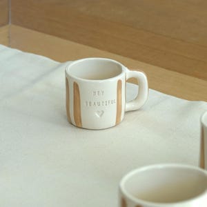 Pode incluir: Uma pequena caneca de cerâmica branca com listras verticais bege e uma alça. A caneca apresenta as palavras "HEY BEAUTIFUL" e um símbolo de coração. A caneca está sobre uma superfície de tecido branco.