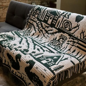 May include: A cream and dark green crocheted blanket with a geometric pattern, draped over a gray sofa. The blanket features a repeating design of stylized shapes and figures, with a fringed edge.