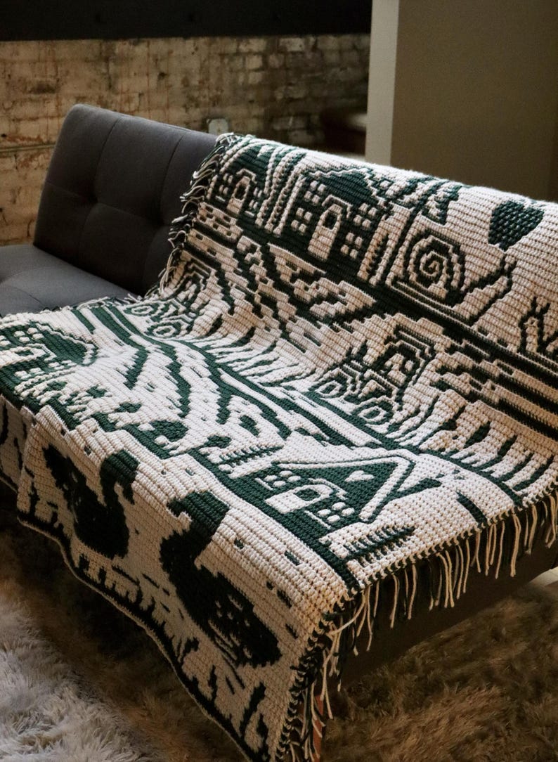 May include: A cream and dark green crocheted blanket with a geometric pattern, draped over a gray sofa. The blanket features a repeating design of stylized shapes and figures, with a fringed edge.
