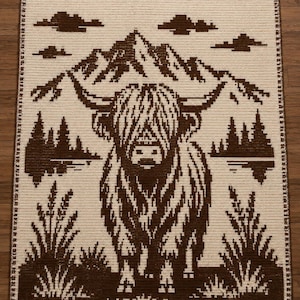 May include: A rectangular crochet blanket featuring a two-tone brown and cream design. The pattern depicts a Highland cow centered in front of a mountain range, pine trees, and clouds, with a decorative border framing the entire piece.