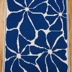 Puede incluir: Una manta rectangular de ganchillo con un patrón floral audaz y a gran escala en azul profundo y blanco roto. El diseño consiste en pétalos de flores estilizados y angulares dispuestos por toda la superficie, con un borde decorativo fino.