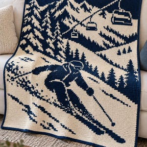 Op de afbeelding: Een marineblauwe en crèmekleurige gehaakte deken met een gedetailleerd grafisch ontwerp van een skiër die een besneeuwde berghelling afdaalt. De scène bevat dennenbomen, een stoeltjeslift met skiliften en bergtoppen.
