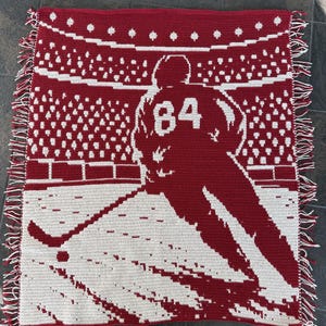 May include: A rectangular crochet blanket featuring a red and white graphic design of a hockey player wearing the number 84 on their jersey. The player is positioned on ice in a stadium setting with a crowd in the background and fringed edges.