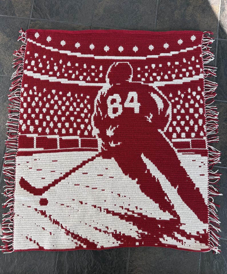 May include: A rectangular crochet blanket featuring a red and white graphic design of a hockey player wearing the number 84 on their jersey. The player is positioned on ice in a stadium setting with a crowd in the background and fringed edges.
