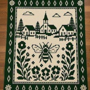 May include: A rectangular crochet blanket featuring a dark green and cream color scheme. The design displays a central bee surrounded by flowers, with a village scene above including a church, houses, trees, and clouds, all framed by a geometric border.
