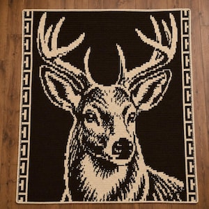 May include: A rectangular crochet blanket featuring a large, detailed deer head graphic in cream against a dark brown background. The design is framed by a cream-colored border with a repeating geometric pattern along the left and right sides.