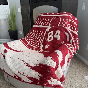 May include: A red and white crocheted blanket featuring a graphic of a hockey player wearing a jersey with the number 84 on the back. The design includes a stadium crowd pattern in the background and a textured fringe along the bottom edge.