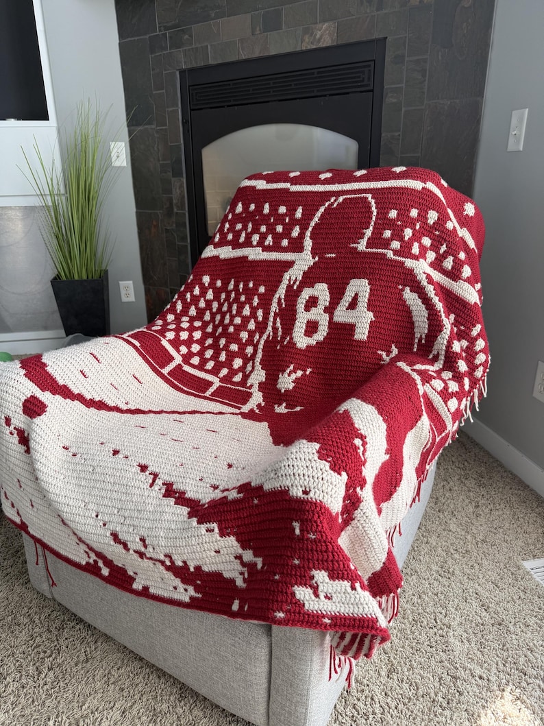 May include: A red and white crocheted blanket featuring a graphic of a hockey player wearing a jersey with the number 84 on the back. The design includes a stadium crowd pattern in the background and a textured fringe along the bottom edge.