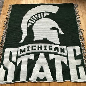 May include: A green and white woven blanket featuring the Michigan State Spartans logo and the words "MICHIGAN STATE". The blanket has a fringe border and is displayed on a wooden floor.