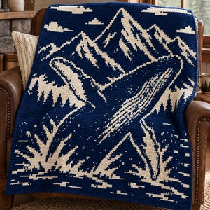 May include: A dark blue and cream crochet blanket featuring a graphic design of a whale breaching out of the water in front of a mountain range. The pattern includes stylised clouds in the sky and waves at the base, created with a pixelated stitch.