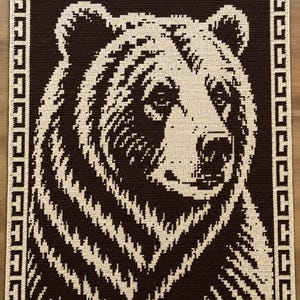 Può includere: Una coperta rettangolare all'uncinetto che presenta un grande e dettagliato ritratto di un orso in filato marrone scuro e crema. L'immagine centrale dell'orso è incorniciata da un bordo verticale sui lati sinistro e destro, con un motivo geometrico greco.