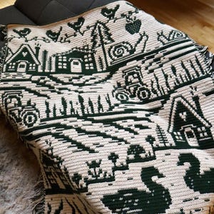 May include: A cream and dark green crocheted blanket featuring a rural landscape design. The blanket depicts houses, tractors, trees, birds, and swans. The blanket is draped over a dark gray couch.