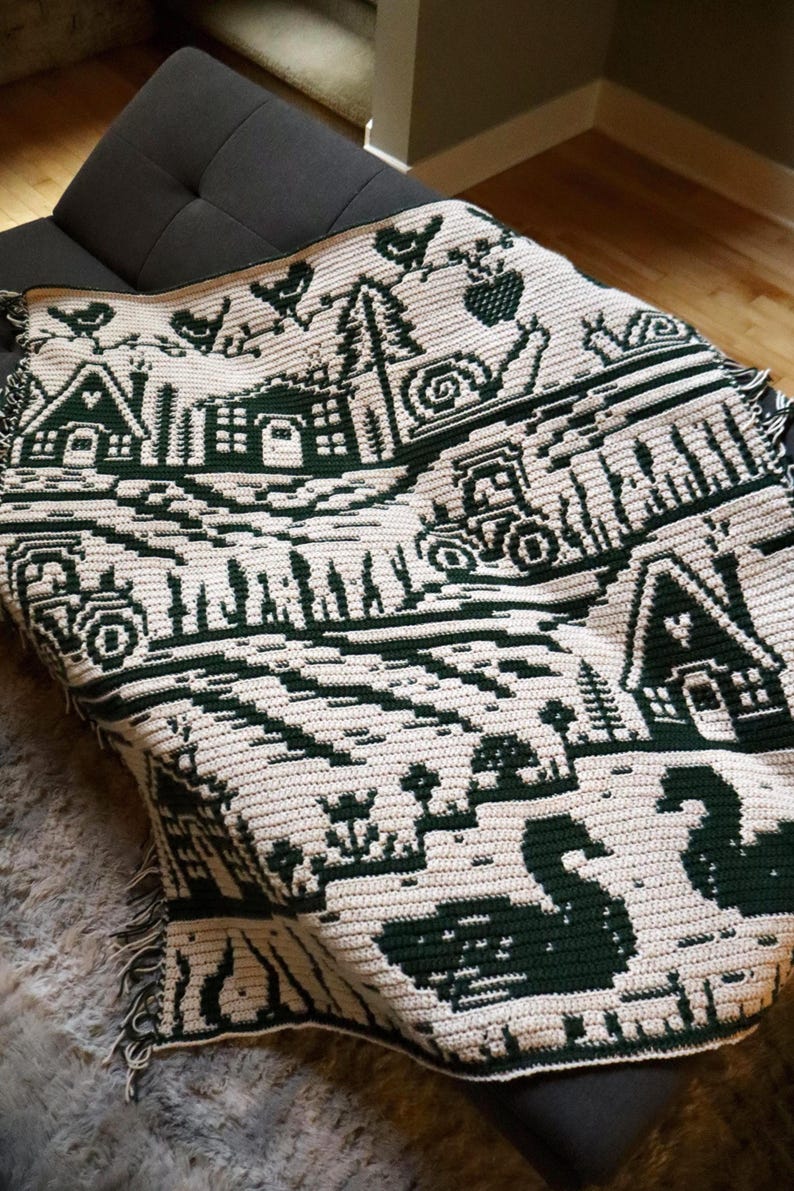 May include: A cream and dark green crocheted blanket featuring a rural landscape design. The blanket depicts houses, tractors, trees, birds, and swans. The blanket is draped over a dark gray couch.