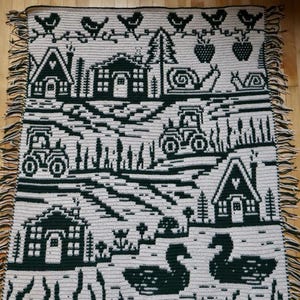 May include: A cream-colored crocheted blanket with a dark green countryside design. The blanket features houses, tractors, birds, strawberries, snails, and swans. The blanket has a fringe border.
