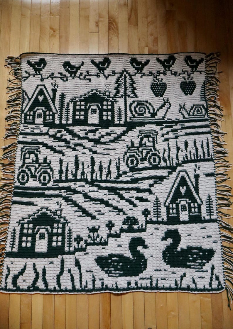 May include: A cream-colored crocheted blanket with a dark green countryside design. The blanket features houses, tractors, birds, strawberries, snails, and swans. The blanket has a fringe border.