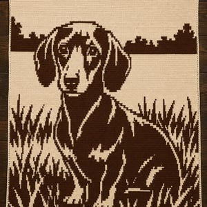 May include: A rectangular crochet blanket featuring a two-tone brown and cream pixelated design of a dachshund sitting in a field of tall grass. The dog is centred, facing forward, with a landscape of trees visible in the background horizon.