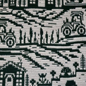 May include: A handmade, crocheted blanket featuring a repeating pattern of dark green rural scenes on a cream background. The design includes houses, trees, tractors, and a swan, creating a cozy, rustic aesthetic. The blanket is likely made of soft yarn.