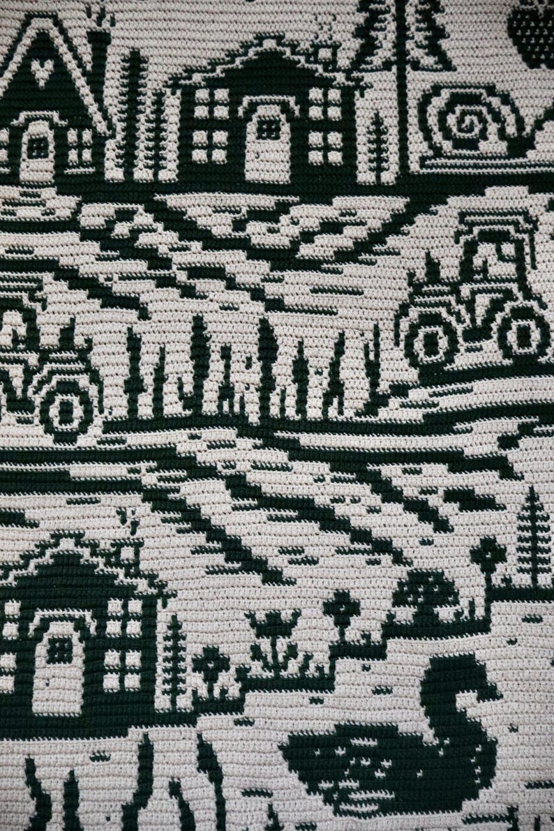 May include: A handmade, crocheted blanket featuring a repeating pattern of dark green rural scenes on a cream background. The design includes houses, trees, tractors, and a swan, creating a cozy, rustic aesthetic. The blanket is likely made of soft yarn.