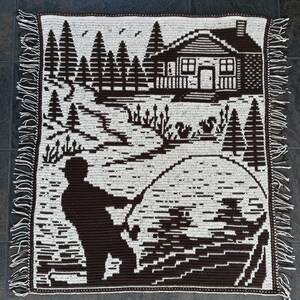 May include: A brown and white crocheted blanket featuring a landscape scene. The design includes a cabin, trees, a fisherman, and a lake. The blanket has a fringed edge.