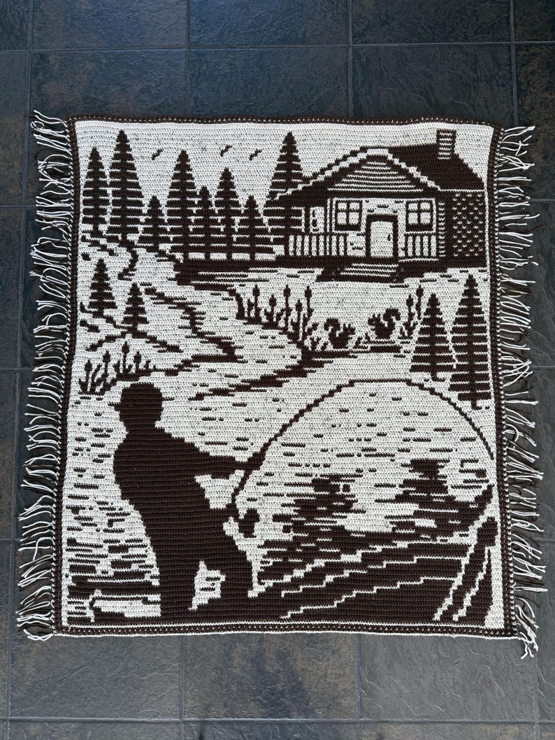 May include: A brown and white crocheted blanket featuring a landscape scene. The design includes a cabin, trees, a fisherman, and a lake. The blanket has a fringed edge.