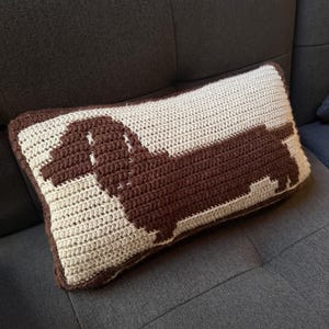 May include: A rectangular crochet pillow featuring a dark brown dachshund silhouette centered against a cream-colored background. The pillow has a dark brown border and a textured yarn construction, showcasing a clear canine shape in profile.