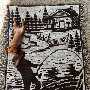 May include: A brown and cream-colored woven throw blanket featuring a cabin scene with trees, a lake, and a fisherman. The blanket has a fringed edge and a dachshund dog is lying on top of it.