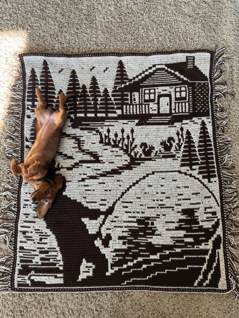 May include: A brown and cream-colored woven throw blanket featuring a cabin scene with trees, a lake, and a fisherman. The blanket has a fringed edge and a dachshund dog is lying on top of it.