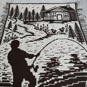May include: A brown and cream-colored crocheted throw blanket featuring a cabin scene with a fisherman. The design includes trees, a cabin, and a fisherman casting a line. The blanket has a fringed edge.