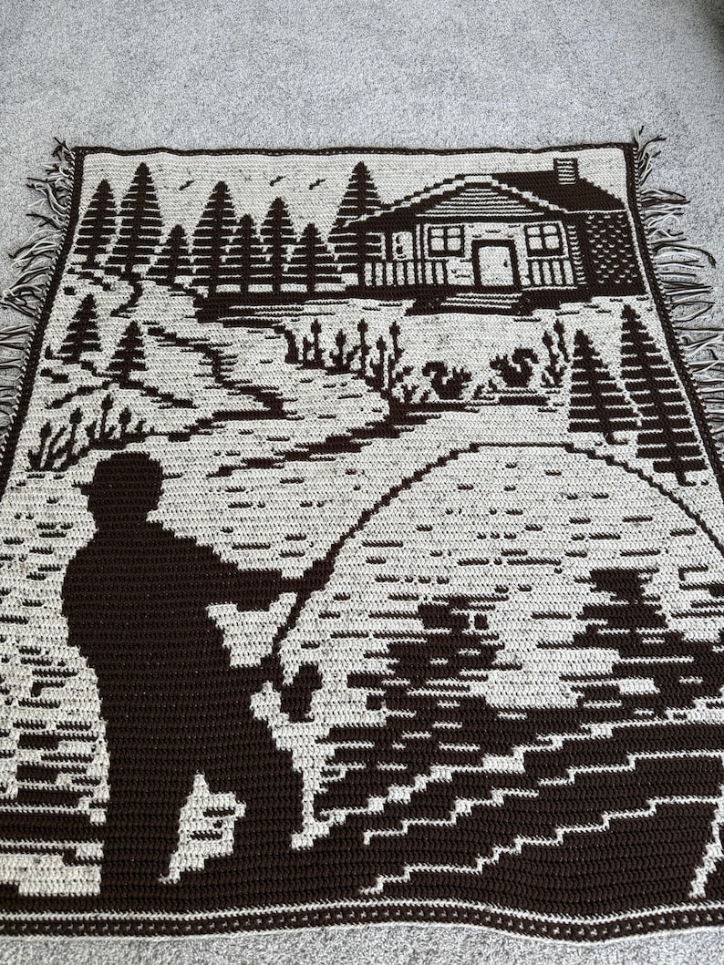 May include: A brown and cream-colored crocheted throw blanket featuring a cabin scene with a fisherman. The design includes trees, a cabin, and a fisherman casting a line. The blanket has a fringed edge.