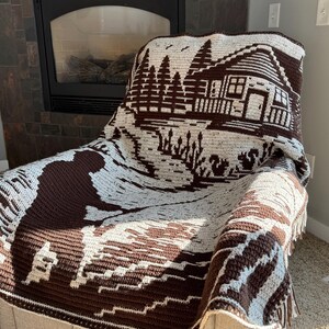 May include: A brown and cream-colored crocheted blanket draped over a chair. The blanket features a detailed landscape design with a cabin, trees, and water. The blanket has a fringed edge and is made of soft yarn.