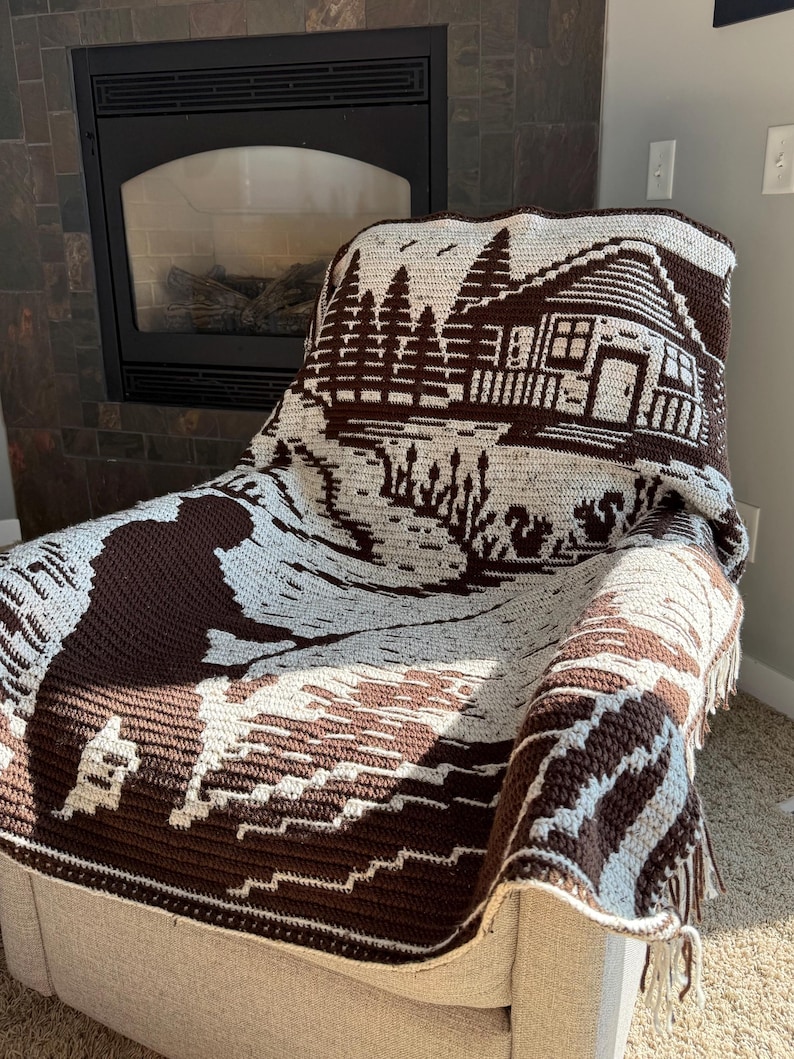 May include: A brown and cream-colored crocheted blanket draped over a chair. The blanket features a detailed landscape design with a cabin, trees, and water. The blanket has a fringed edge and is made of soft yarn.