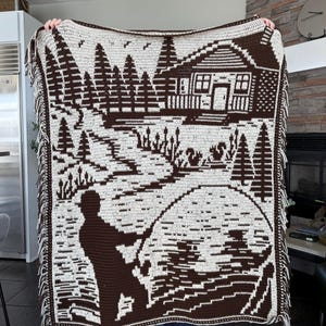 May include: A brown and white crocheted blanket featuring a fishing scene. The design includes a cabin, trees, a fisherman, and a lake. The blanket has a fringed edge and is held up by a person.