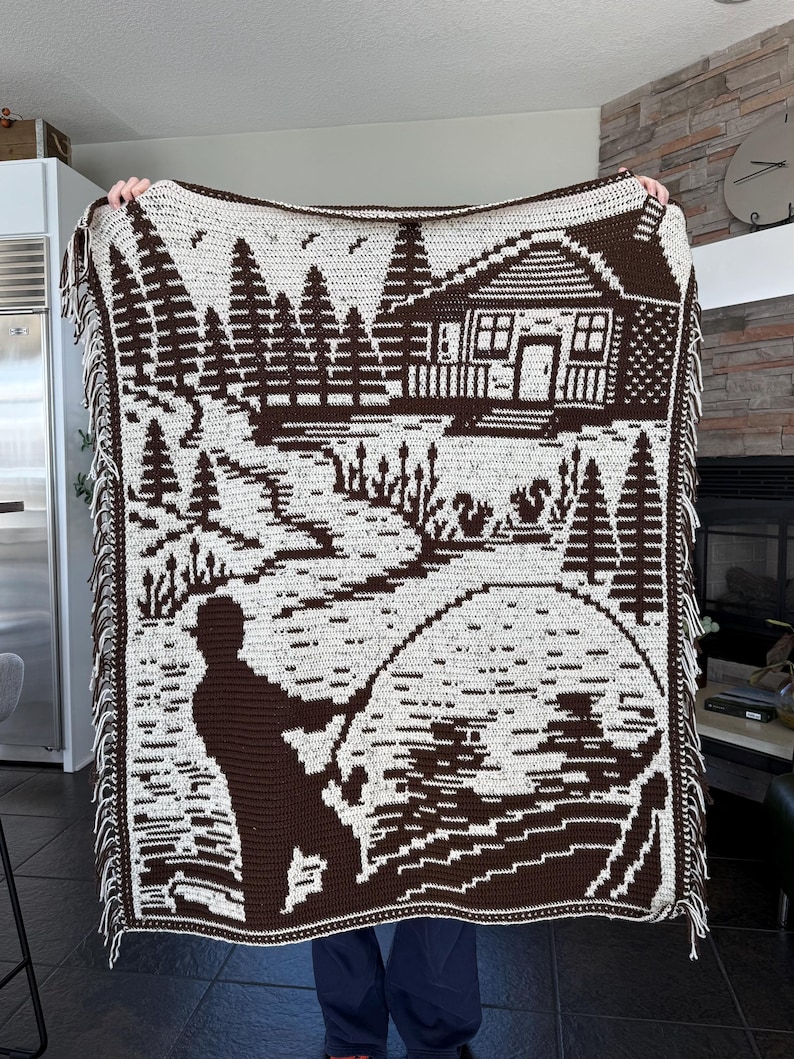 May include: A brown and white crocheted blanket featuring a fishing scene. The design includes a cabin, trees, a fisherman, and a lake. The blanket has a fringed edge and is held up by a person.