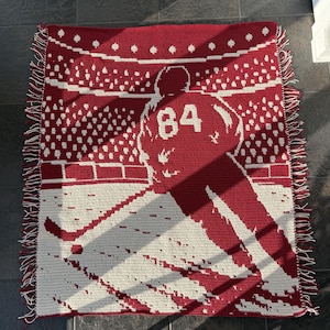 Peut inclure: Une couverture en crochet rectangulaire présentant un motif bicolore rouge et crème. Le dessin représente un joueur de hockey portant un maillot avec le numéro 84 au dos, sur une patinoire devant une foule. Les bords sont dotés de franges.