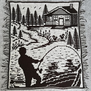 May include: A crocheted throw blanket featuring a black and white landscape scene. The design depicts a fisherman, a cabin, trees, and a body of water. The blanket has a fringed edge.