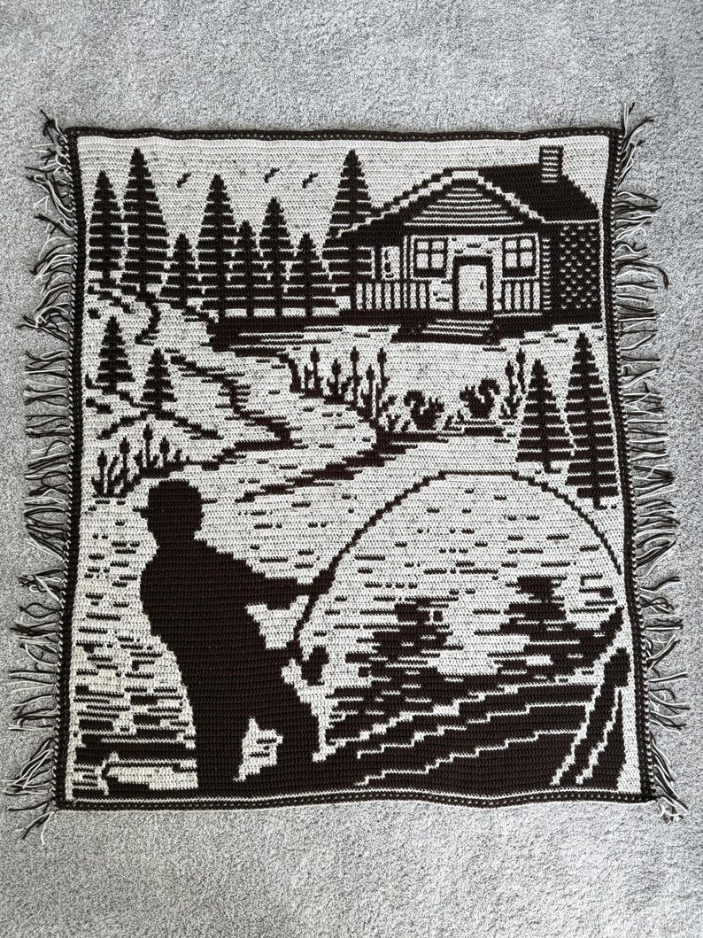 May include: A crocheted throw blanket featuring a black and white landscape scene. The design depicts a fisherman, a cabin, trees, and a body of water. The blanket has a fringed edge.