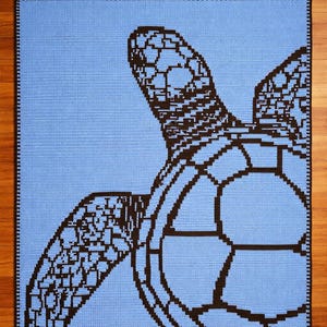 Puede incluir: Una manta de ganchillo con un diseño de tortuga marina. La tortuga es de color marrón oscuro sobre un fondo azul claro. La manta tiene un borde negro. El diseño es probablemente hecho a mano.