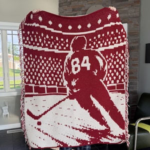 May include: A rectangular crochet blanket featuring a two-tone red and white design. The pattern depicts a hockey player wearing jersey number 84 on the ice, with a stadium crowd in the background. The blanket is finished with a fringed edge.