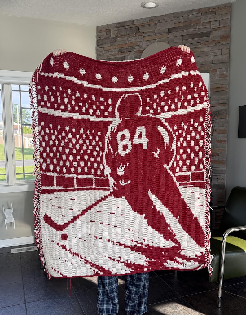 May include: A rectangular crochet blanket featuring a two-tone red and white design. The pattern depicts a hockey player wearing jersey number 84 on the ice, with a stadium crowd in the background. The blanket is finished with a fringed edge.