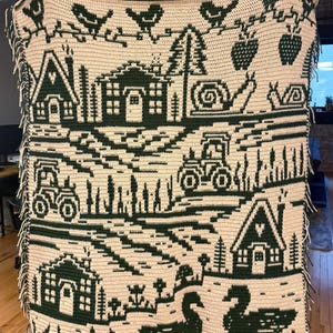 May include: A cream-colored, crocheted blanket with a dark green countryside design. The blanket features houses, tractors, birds, snails, and swans. The edges have a fringe. The blanket is held up by a person.