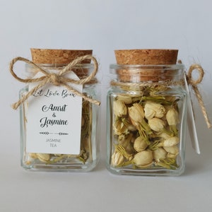 Puede incluir: Dos pequeños frascos de vidrio con tapas de corcho, llenos de flores de jazmín secas. Cada frasco tiene un lazo de cuerda y una etiqueta blanca con el texto "Lit Love Brew Amrit & Jasmine JASMINE TEA".