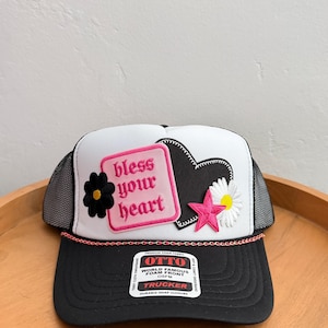 Könnte beinhalten: Eine weiß-schwarze Trucker-Cap mit einem rosafarbenen rechteckigen Aufnäher mit dem Text bless your heart, einer schwarzen Blume, einer schwarzen Herzumrandung, einem weißen Gänseblümchen und einem rosafarbenen Stern. Eine dünne rosa Kette verziert den schwarzen Schirm.