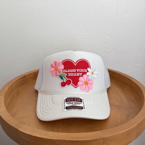 May include: White trucker hat with a mesh back and a curved brim. The front features a red heart with the words "Bless Your Heart" and floral and cherry embellishments. A tag on the brim reads "Otto World Famous Foam Front Trucker".