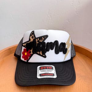 Może przedstawiać: Biało-czarna czapka typu trucker z naszywką w kształcie gwiazdy w panterkę, czerwonym kwiatem i napisem Mama czcionką w kolorze czarnym. Po prawej stronie znajduje się złota błyskawica. Na daszku widnieje naklejka OTTO World Famous Foam Front.