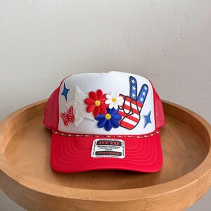 May include: Red and white trucker hat with a peace sign, stars, and flowers. The hat features a red brim and mesh back, with the text "OTTO" on a tag.